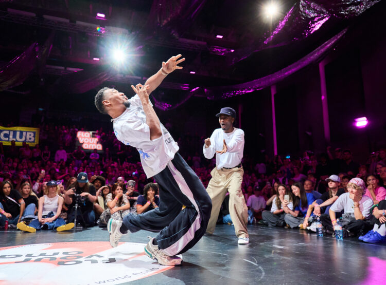 Zwei Männer breakdancen auf der Bühne vor einer jubelnden Menge und bunten Lichtern im Hintergrund.