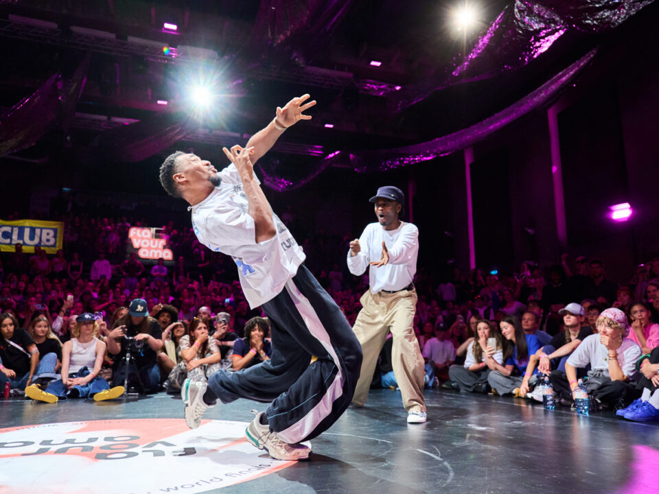Zwei Männer breakdancen auf der Bühne vor einer jubelnden Menge und bunten Lichtern im Hintergrund.