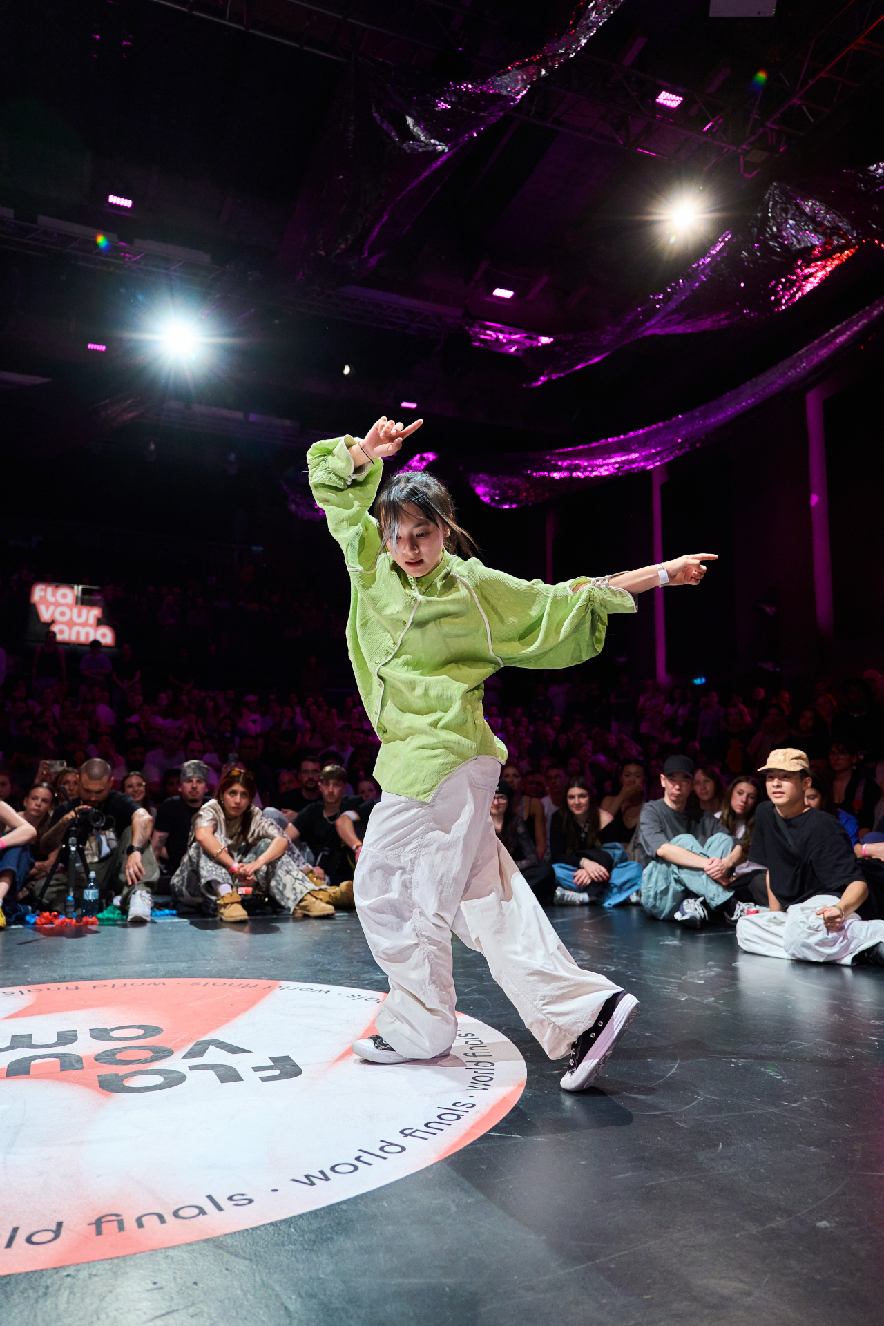 Ein T&auml;nzer in gr&uuml;nem Hemd und wei&szlig;er Hose tritt auf der B&uuml;hne auf, umgeben von einem Publikum bei einem Tanzwettbewerb.