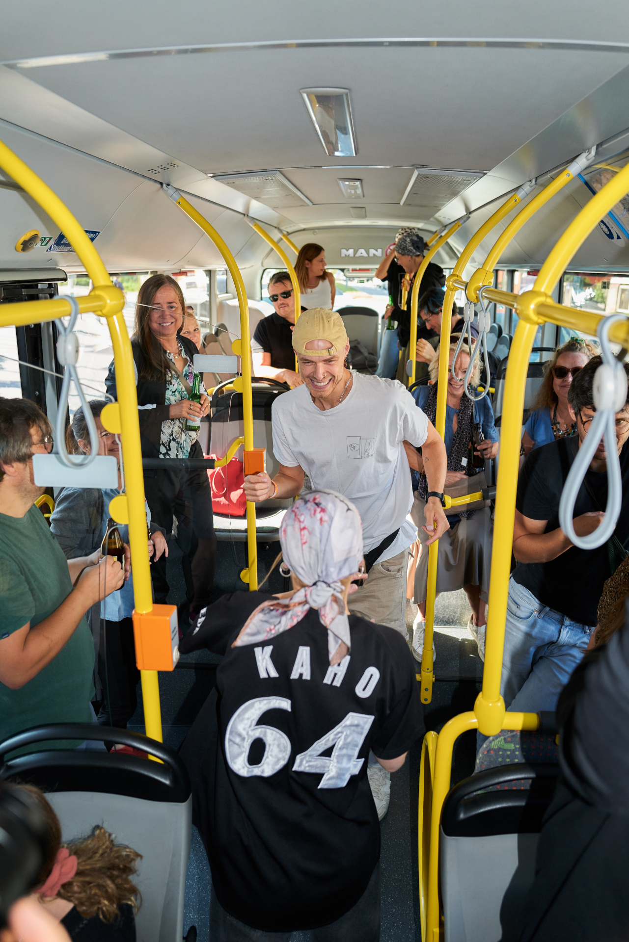 Menschen l&auml;cheln und unterhalten sich in einem &uuml;berf&uuml;llten Stadtbus, einige halten sich an gelben Stangen fest und machen Fotos.