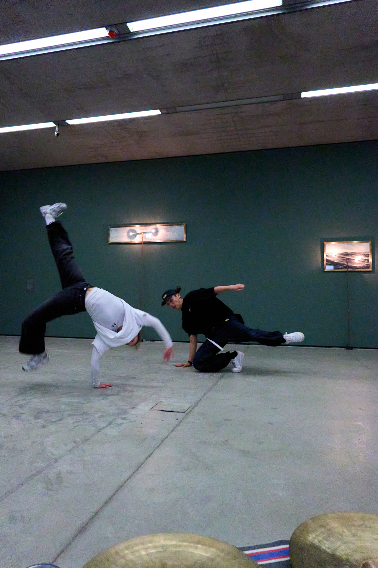 Zwei Personen, die drinnen auf einem glatten Boden breakdancen, mit gr&uuml;nen W&auml;nden und Kunstwerken im Hintergrund.