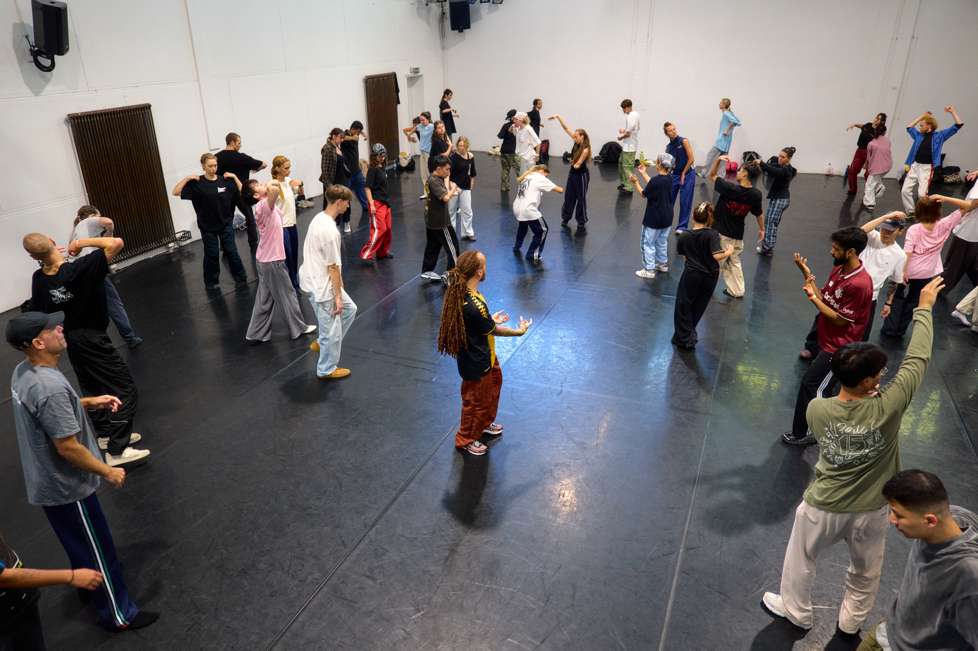 Eine große Gruppe von Menschen übt gemeinsam Tanzschritte in einem geräumigen Studio mit schwarzem Boden und weißen Wänden.