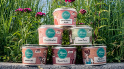 Dosen mit Tierfutter, die im Freien vor hohem Gras und rosa Blumen unter blauem Himmel gestapelt sind.