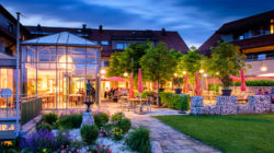 Restaurantterrasse mit Tischen, Sonnenschirmen und Garten, in der Abenddämmerung von warmen Lichtern beleuchtet.
