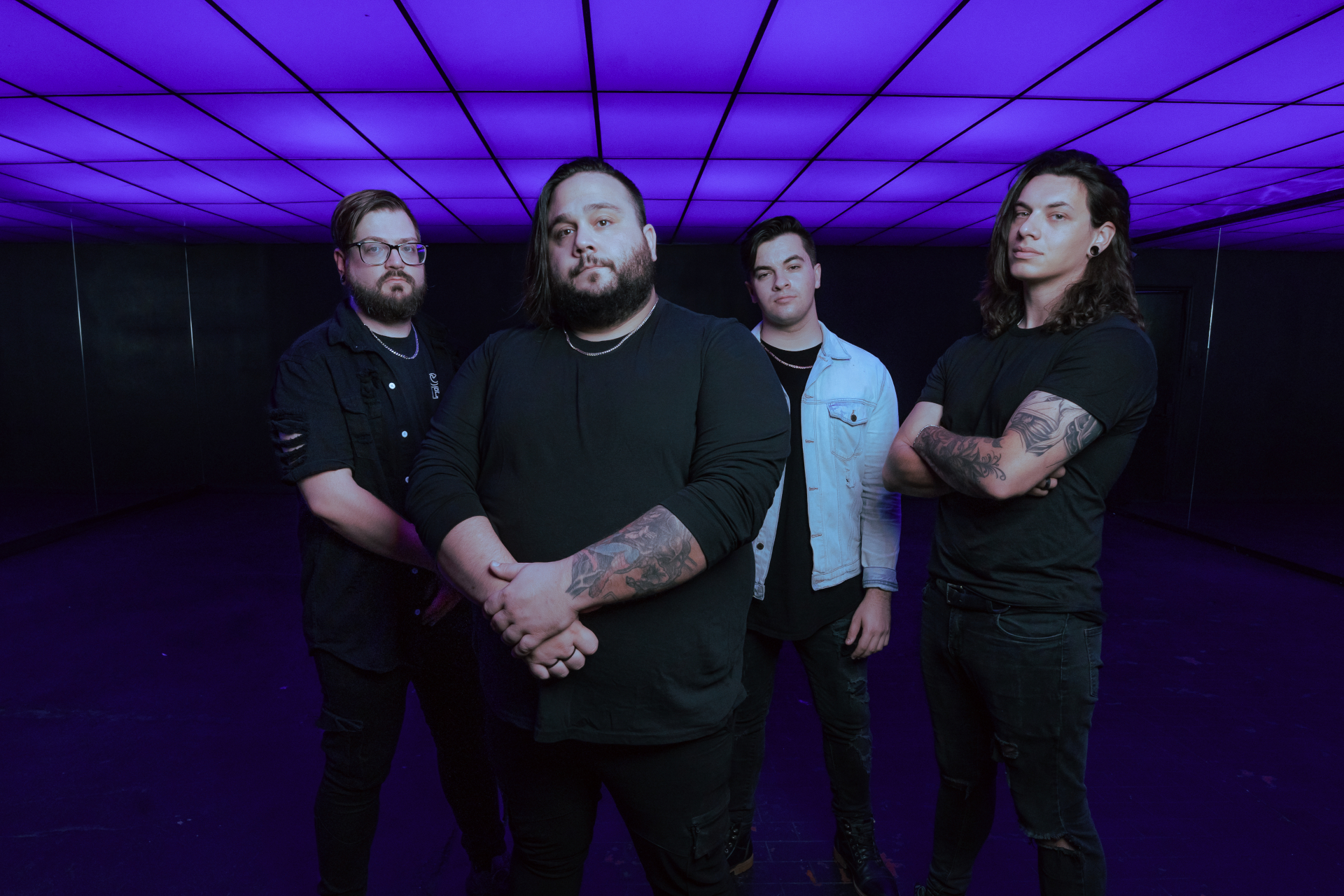 Vier Männer mit Tätowierungen posieren unter einer lila beleuchteten Decke, selbstbewusst in die Kamera blickend.