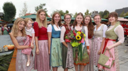 Eine Gruppe von Frauen und ein junges Mädchen in traditioneller österreichischer Kleidung lächeln im Freien, eine hält einen Blumenstrauß.