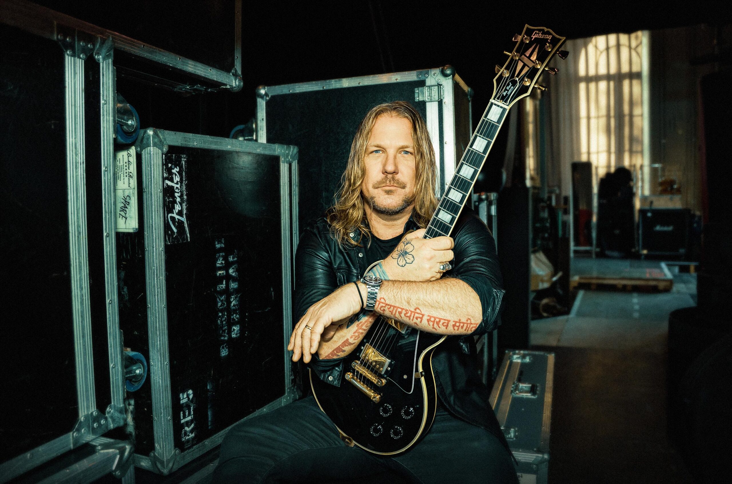 Mann mit langem blondem Haar, der eine schwarze E-Gitarre in der Hand hält und zwischen Gerätekoffern in einem Backstage-Bereich sitzt.