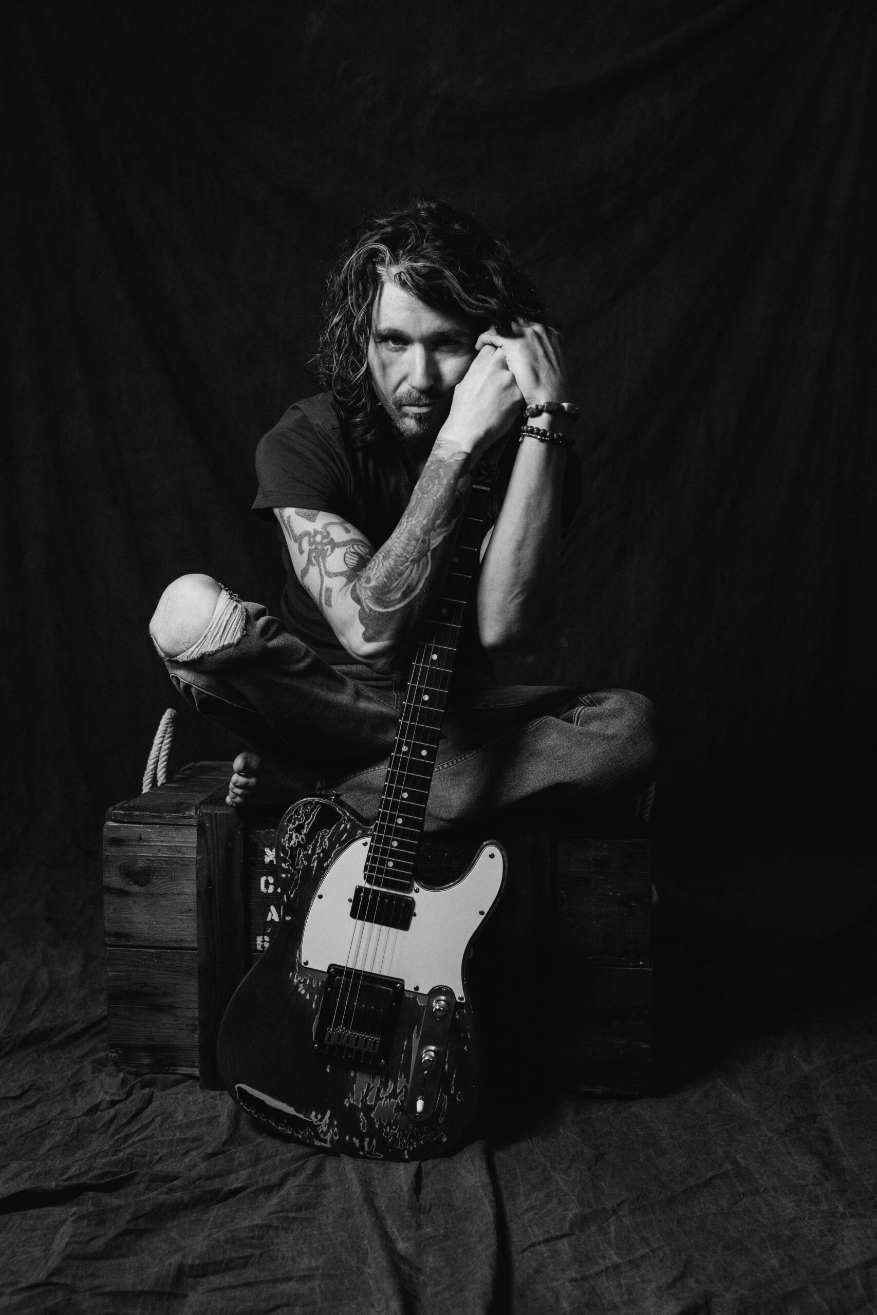 Schwarz-Weiß-Foto eines sitzenden Mannes, der eine Gitarre hält, mit nachdenklichem Gesichtsausdruck und dunklem Hintergrund.