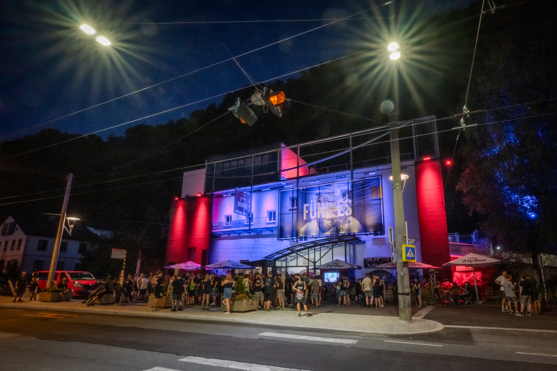 Eine Menschenmenge versammelt sich nachts vor einem hell erleuchteten Gebäude, das von Straßenlaternen angestrahlt wird.