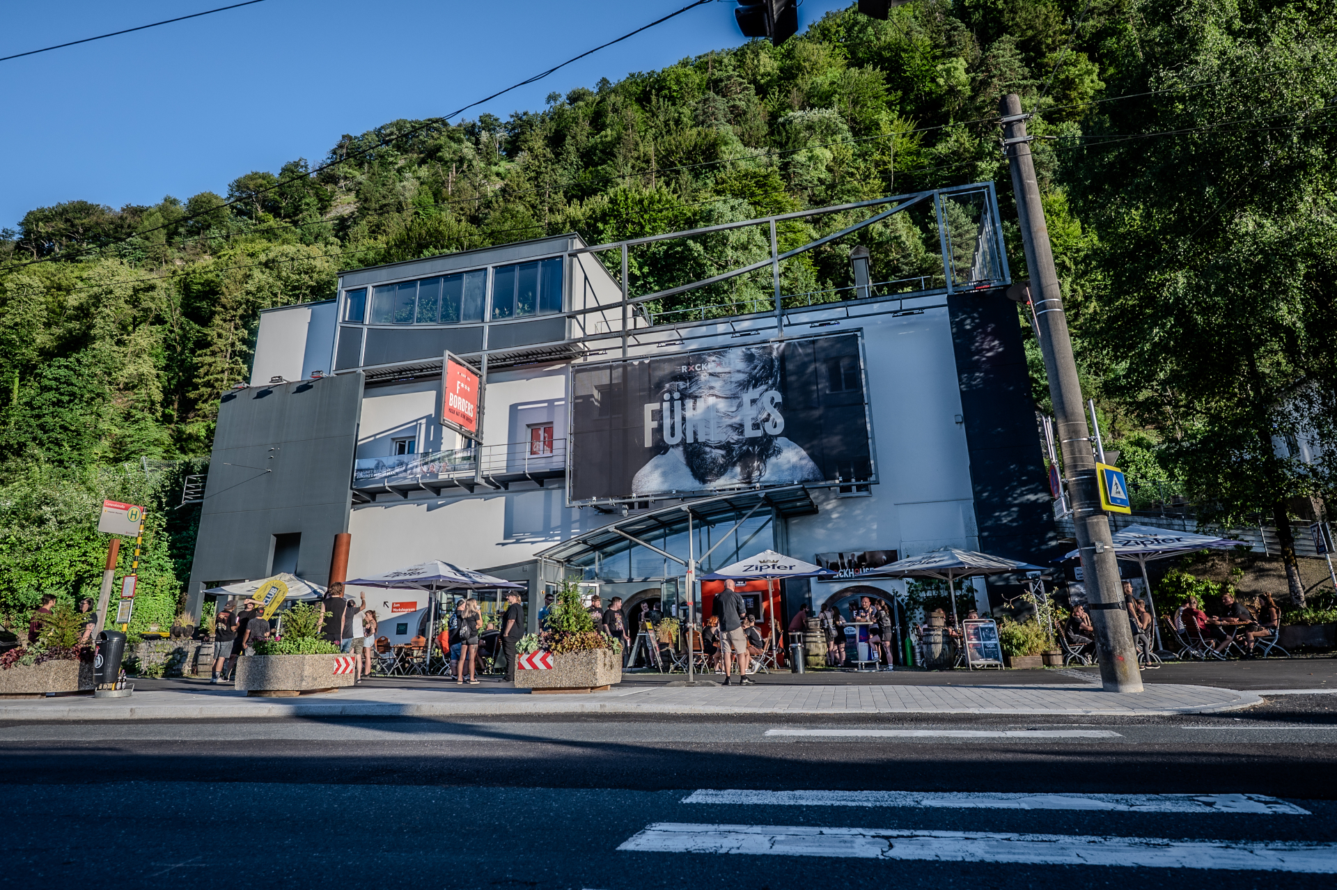 Ein modernes Gebäude mit Sitzgelegenheiten im Freien steht an einer Straße, inmitten von Bäumen und Grünanlagen.
