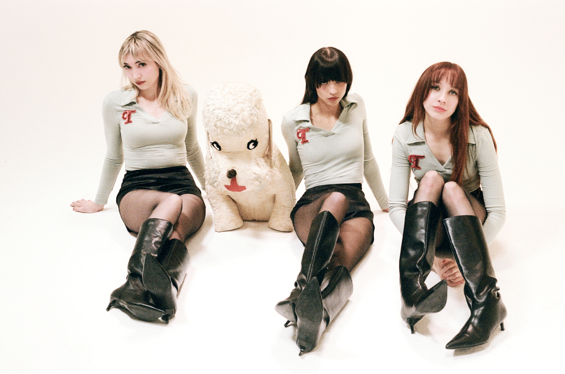 Drei Frauen in passender Kleidung sitzen mit einem großen weißen Pudel-Plüschtier in einem weißen Studio auf dem Boden.