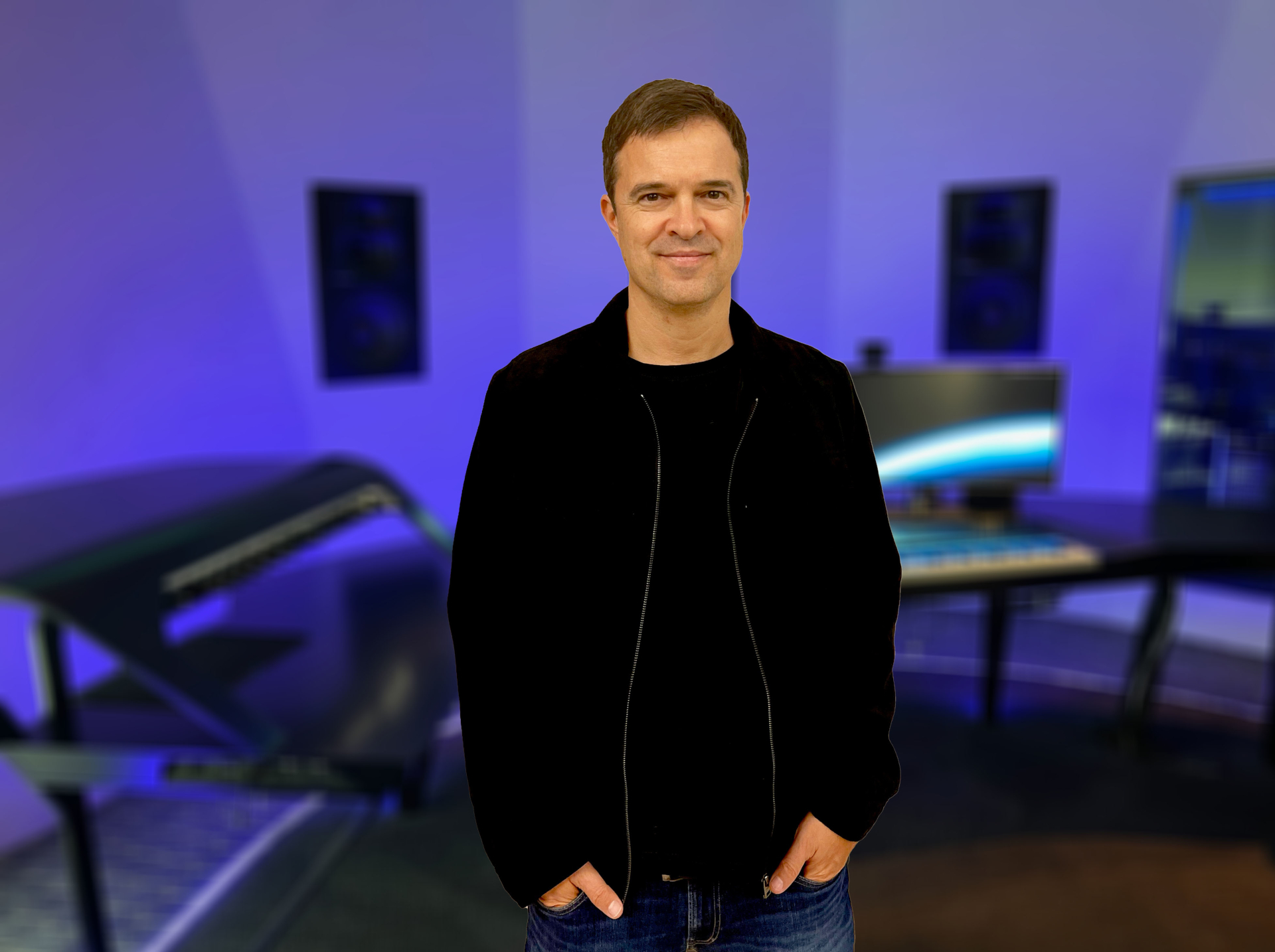 Ein Mann in Freizeitkleidung steht in einem modernen, blau beleuchteten Musikstudio mit Keyboards und Monitoren.