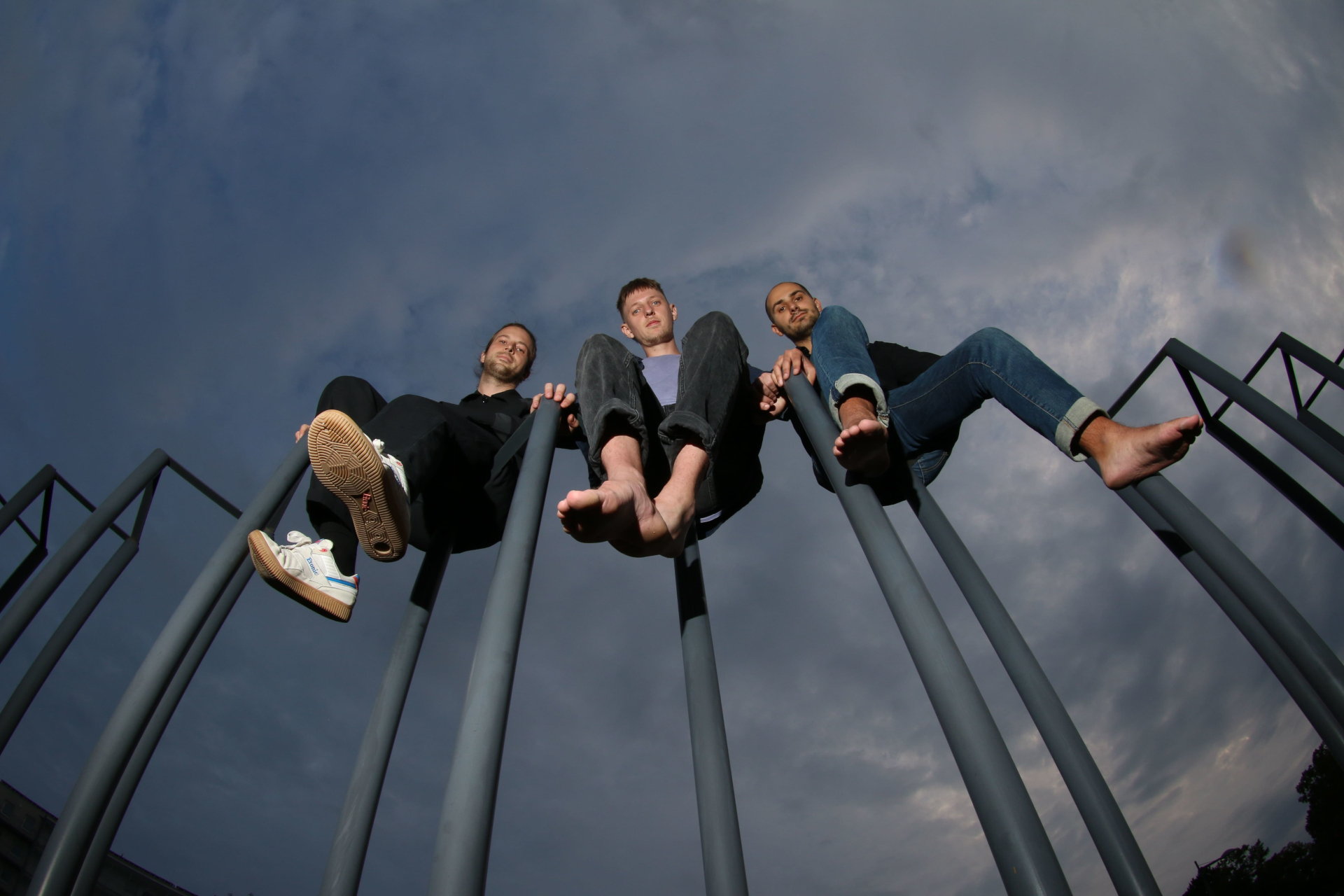 Drei Personen sitzen barfuß auf Metallstangen vor einem wolkenverhangenen Himmel, gesehen aus einer niedrigen Perspektive.