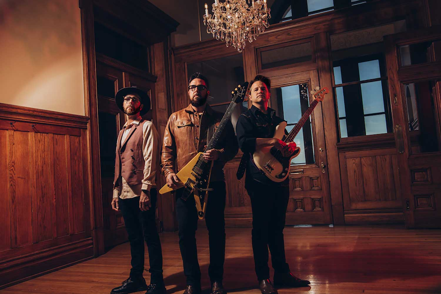 Drei Männer mit Gitarren stehen in einem Holzzimmer unter einem Kronleuchter und posieren für ein Bandfoto.