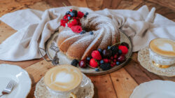 Gugelhupf mit Puderzucker und Beeren, dazu zwei Lattes und zwei Teller auf einem Holztisch.