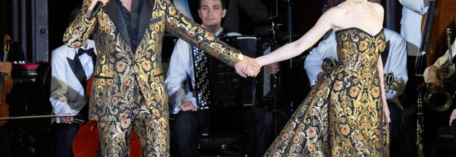 Ein Mann und eine Frau in passender, verzierter Kleidung halten sich auf der Bühne an den Händen, während hinter ihnen Musiker auftreten.