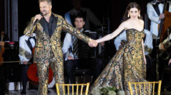 Ein Mann und eine Frau in passender, verzierter Kleidung halten sich auf der Bühne an den Händen, während hinter ihnen Musiker auftreten.