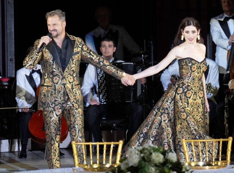 Ein Mann und eine Frau in passender, verzierter Kleidung halten sich auf der Bühne an den Händen, während hinter ihnen Musiker auftreten.