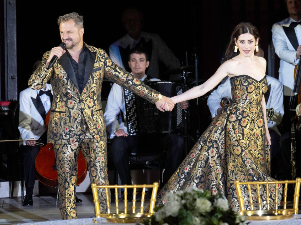 Ein Mann und eine Frau in passender, verzierter Kleidung halten sich auf der Bühne an den Händen, während hinter ihnen Musiker auftreten.