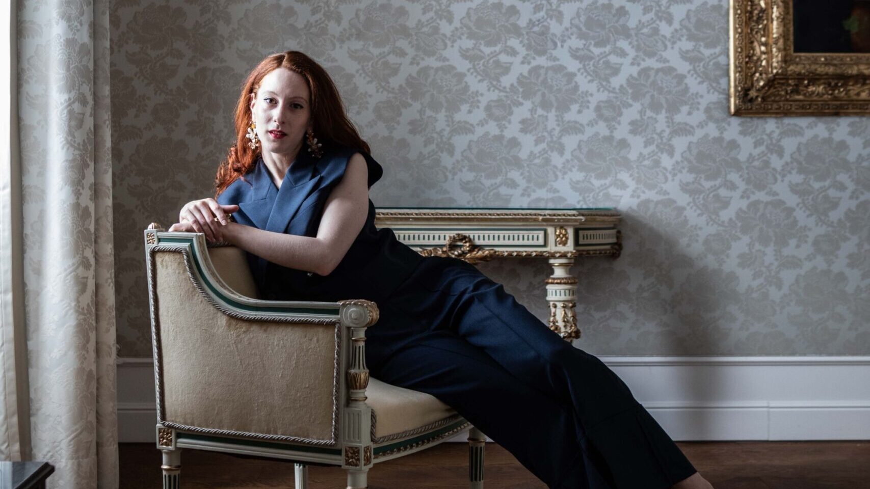 Eine dunkel gekleidete Frau lehnt sich seitlich auf einem verzierten Stuhl in einem stilvollen, sanft beleuchteten Raum zurück.