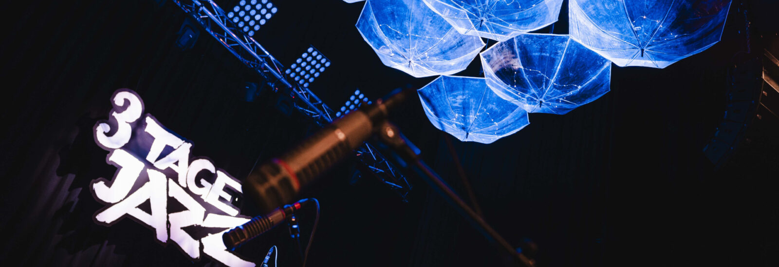 Bühne mit 3 Tage Jazz-Schild, Mikrofonen und blau beleuchteten Schirmen, die von der Decke hängen.