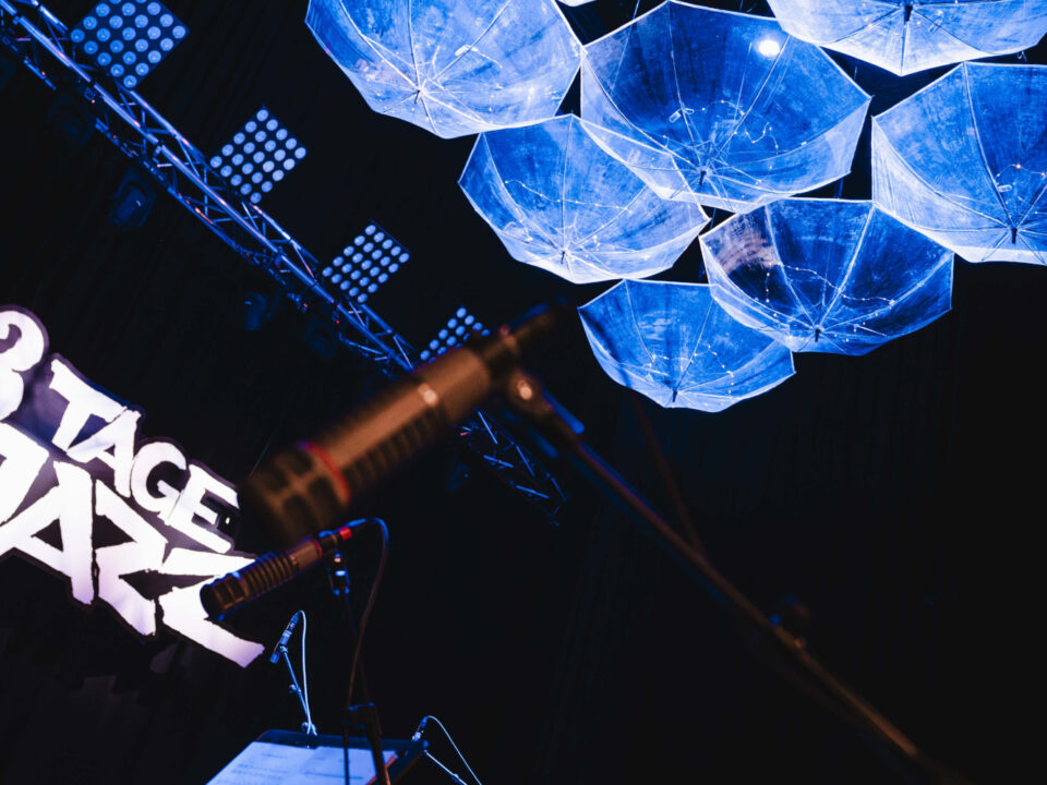 Bühne mit 3 Tage Jazz-Schild, Mikrofonen und blau beleuchteten Schirmen, die von der Decke hängen.