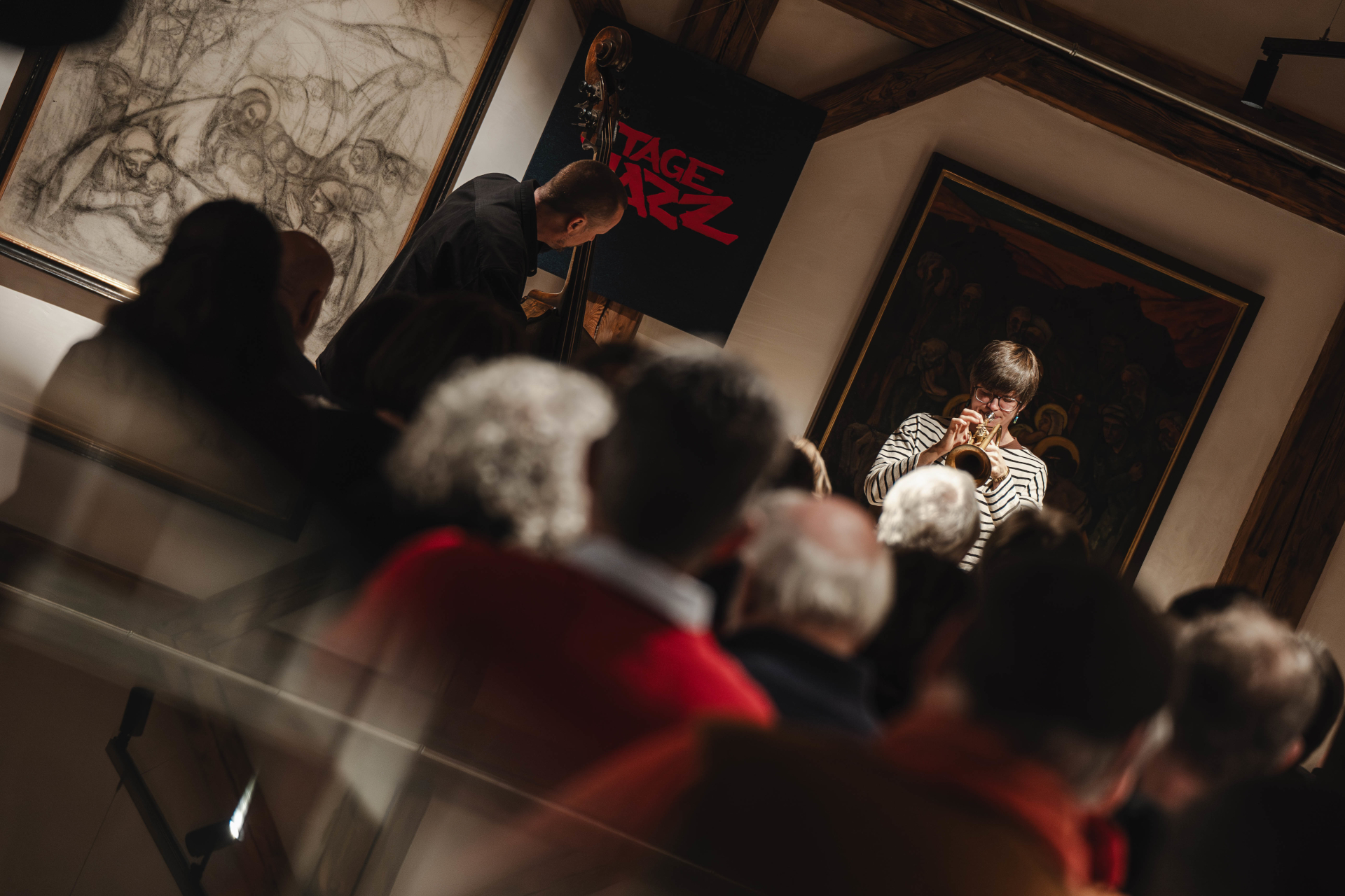 Ein Jazzmusiker spielt auf der B&uuml;hne Trompete vor einem aufmerksamen Publikum in einem kunstvollen, schwach beleuchteten Raum.