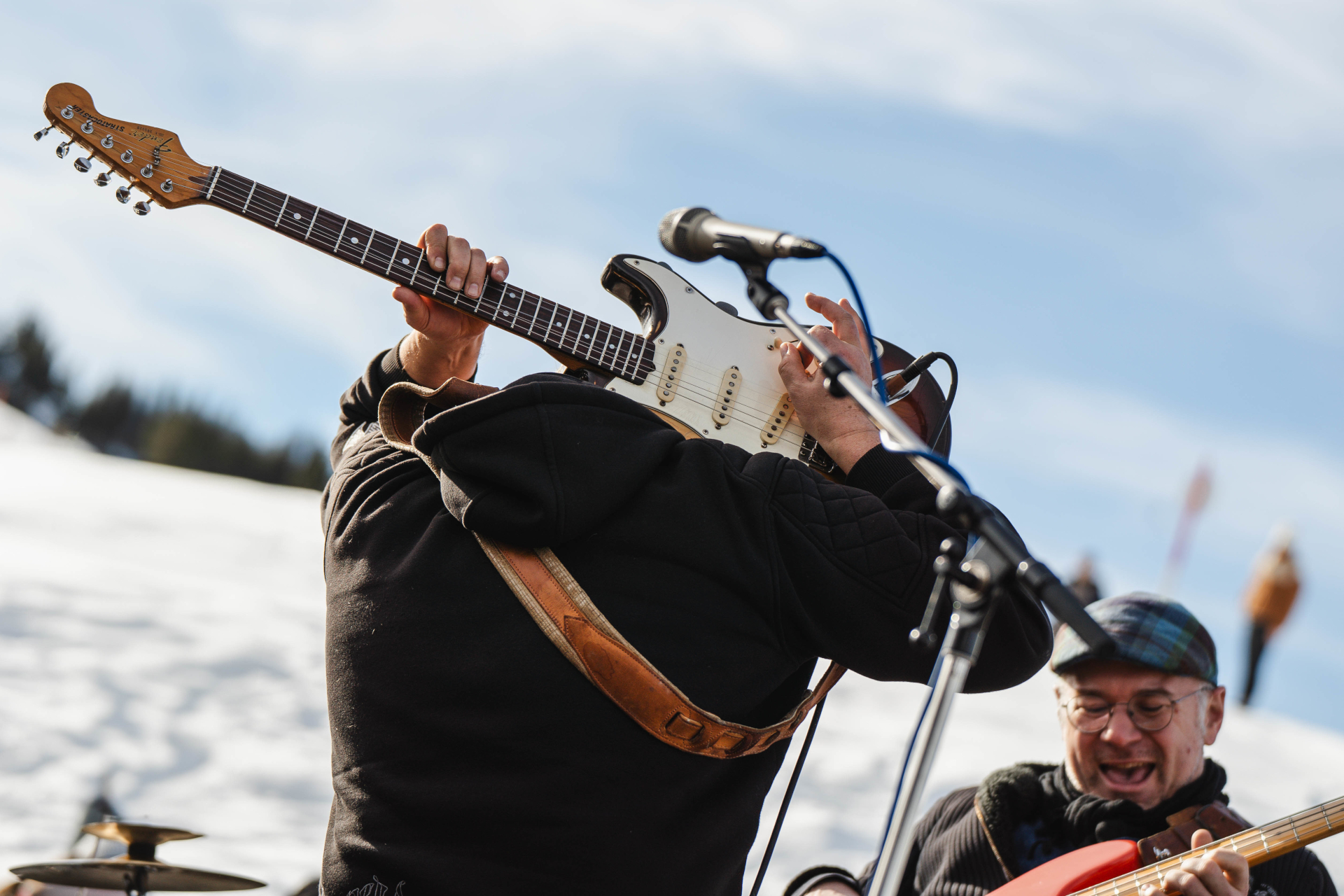 Musiker, der hinter seinem Kopf eine E-Gitarre spielt, im Freien, mit Schnee und einem anderen l&auml;chelnden Musiker in der N&auml;he.