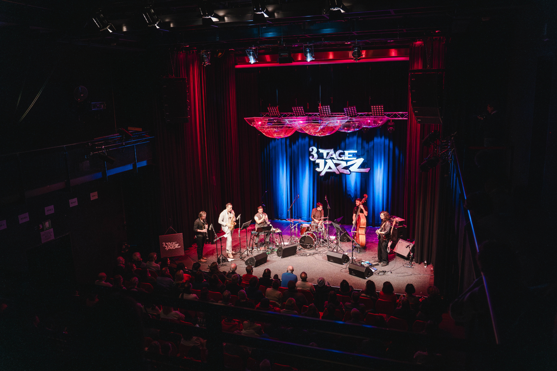 Eine Jazzband tritt auf der B&uuml;hne unter blauem Licht auf, das Publikum sitzt in einem Theater.