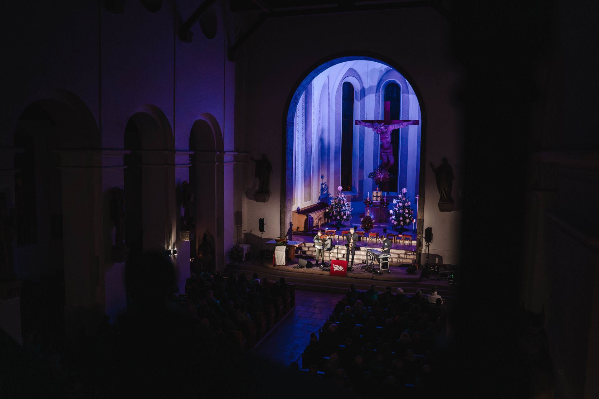 Eine Band spielt auf der B&uuml;hne in einer schwach beleuchteten Kirche mit blauer Beleuchtung und Weihnachtsdekoration.