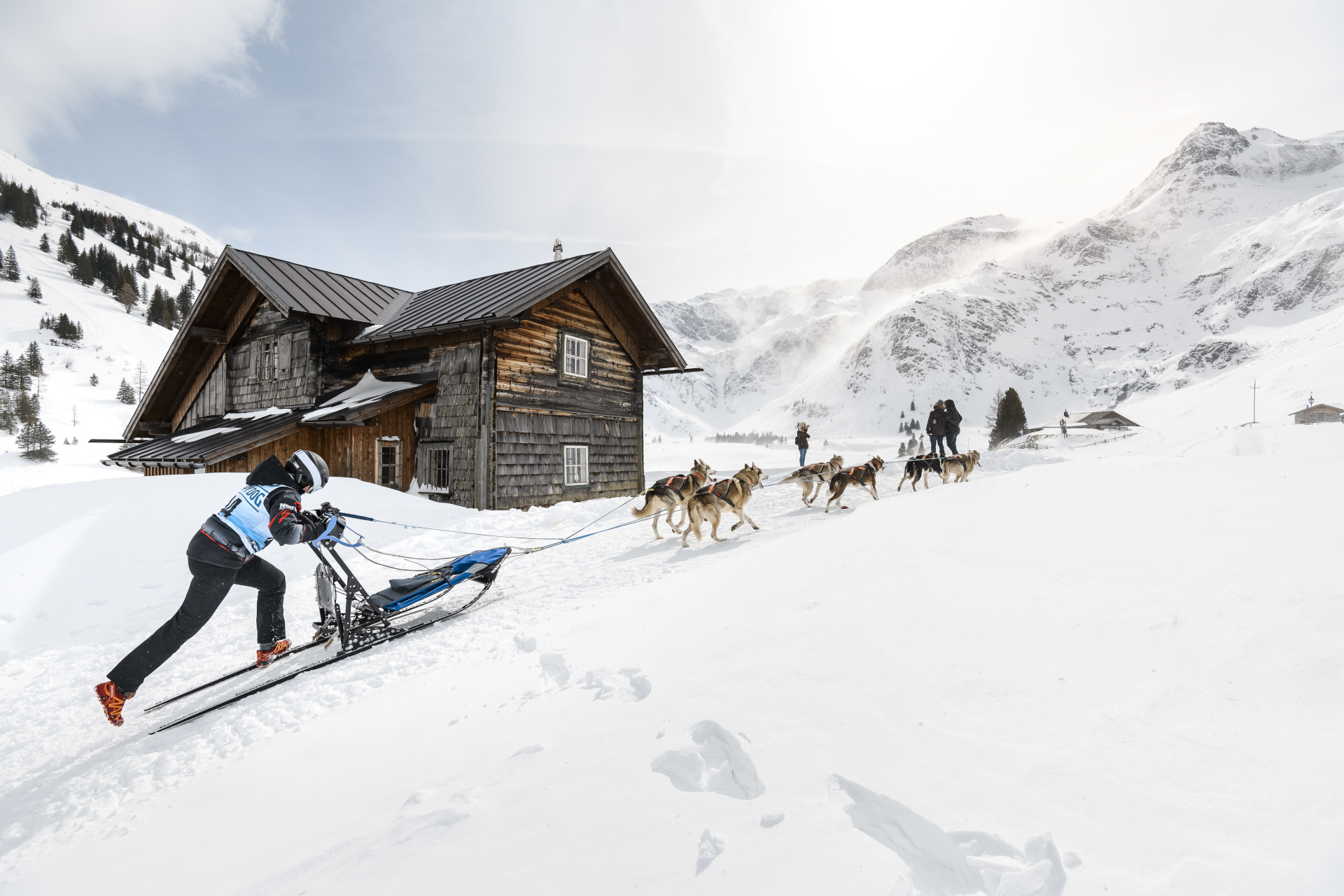 Eine Person auf einem Schlitten wird von Huskys an einer Holzhütte in einer verschneiten Berglandschaft vorbeigezogen.