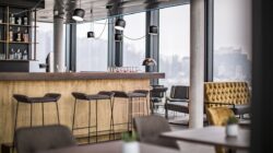 Modernes Café-Interieur mit Barhockern, gelbem Tuftsofa und Pendelleuchten in der Nähe großer Fenster.