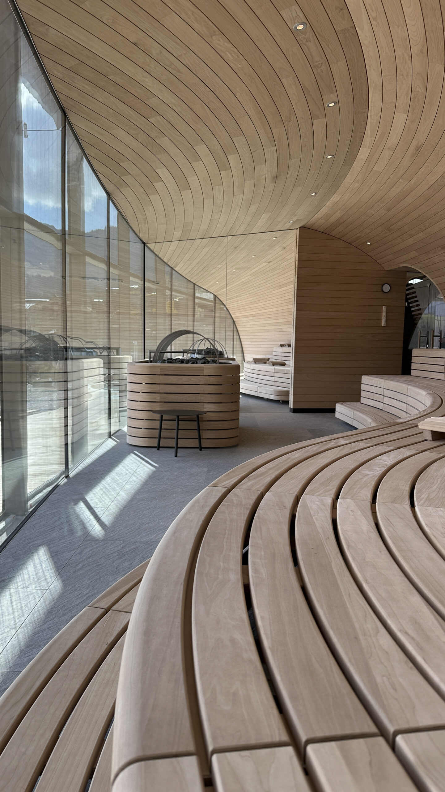 Modernes Interieur mit geschwungenen Holzbänken und -wänden, großen Fenstern und natürlichem Sonnenlicht, das in den Raum strömt.