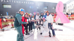 Eine Gruppe von Skifahrern posiert mit Getränken vor einer Skihütte und einer großen rosa Schleife im Schnee.