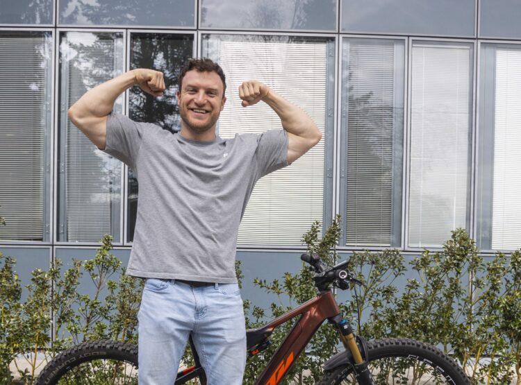 Sascha Huber, Fitness-Influencer aus dem SalzburgerLand, biegt sich und lächelt bei einem Fahrrad und großen Fenstern.