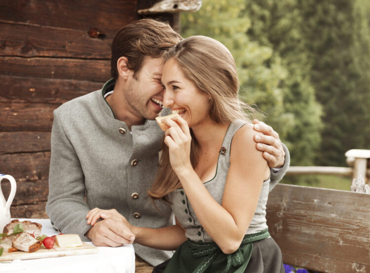 Ein lächelndes Paar sitzt dicht beieinander und teilt sich das Essen vor einer rustikalen Holzhütte, die von viel Grün umgeben ist.