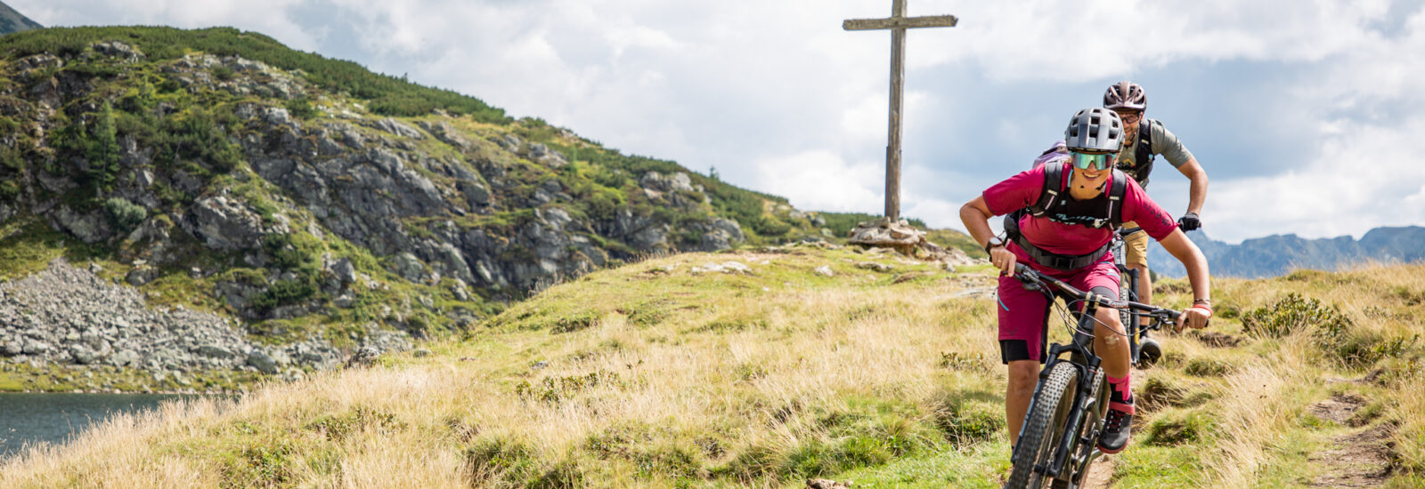2 Mountainbiker, die im Bereich der Oberhütte in Obertauern auf dem Trail mit dem Mountainbike unterwegs sind. Im Hintergrund ein Teil des Oberhüttensees, die Berge und ein Gipfelkreuz.