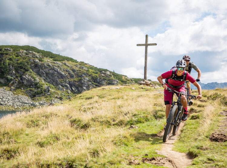2 Mountainbiker, die im Bereich der Oberhütte in Obertauern auf dem Trail mit dem Mountainbike unterwegs sind. Im Hintergrund ein Teil des Oberhüttensees, die Berge und ein Gipfelkreuz.