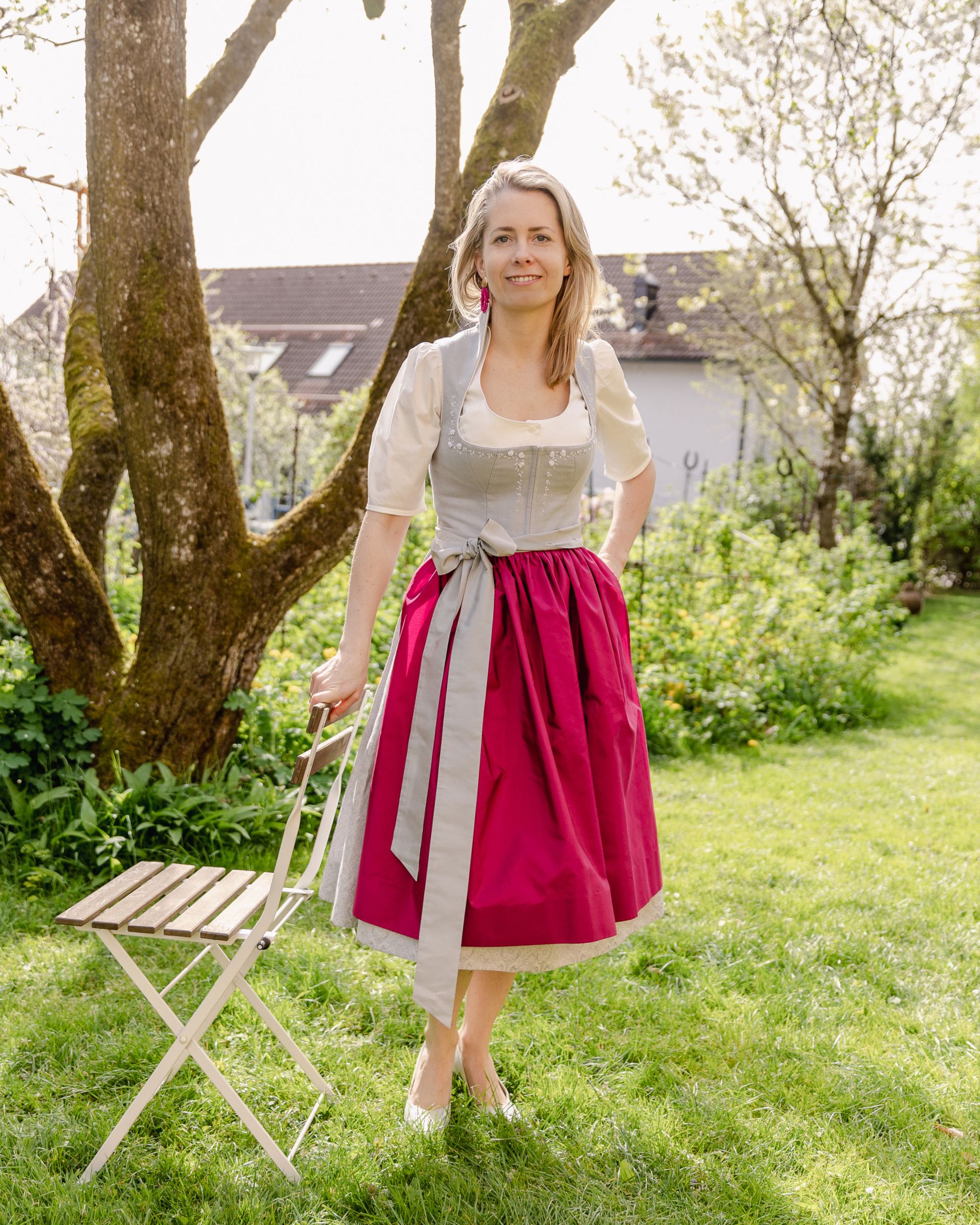 Eine Frau in einem traditionellen österreichischen Dirndlkleid steht in einem sonnigen Garten und hält einen weißen Holzstuhl.