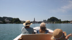 Drei Personen mit Sonnenhüten fahren in einem Boot auf einem See in Richtung einer Stadt mit einem Kirchturm in der Ferne.