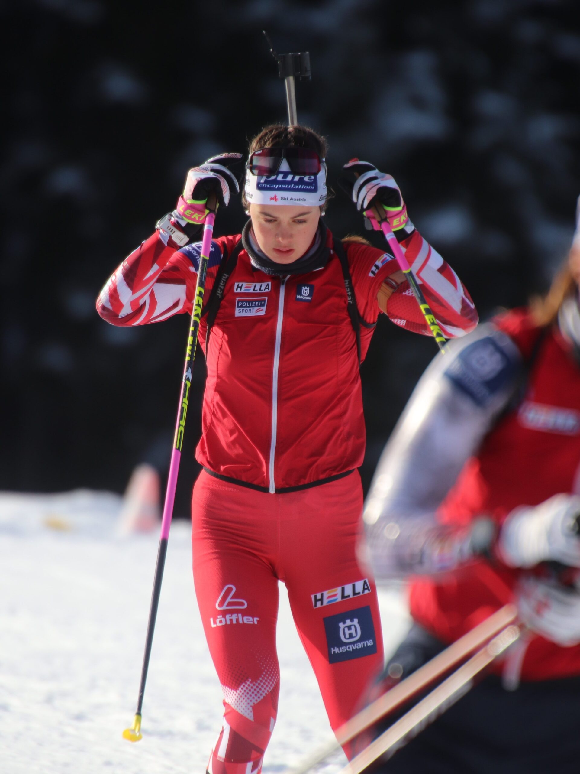 Eine Biathletin in roter Kleidung passt ihr Stirnband an, während sie auf einer verschneiten Strecke Ski fährt.