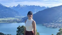 Frau mit Sonnenbrille und Mütze lächelt vor einem See und Bergen an einem sonnigen Tag.