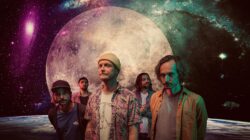 Fünf Männer stehen vor einem großen Mond- und Sternenhintergrund, der eine surreale, kosmische Stimmung erzeugt.