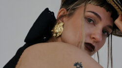 Frau mit dunklem Lippenstift, goldenen Ohrringen und Hut; Hahntattoo auf dem Arm; Blick in die Kamera.