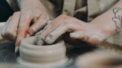 Nahaufnahme von Händen, die nassen Ton auf einer Töpferscheibe formen - eine kreative Auszeit mit sichtbaren Tattoos an den Handgelenken.