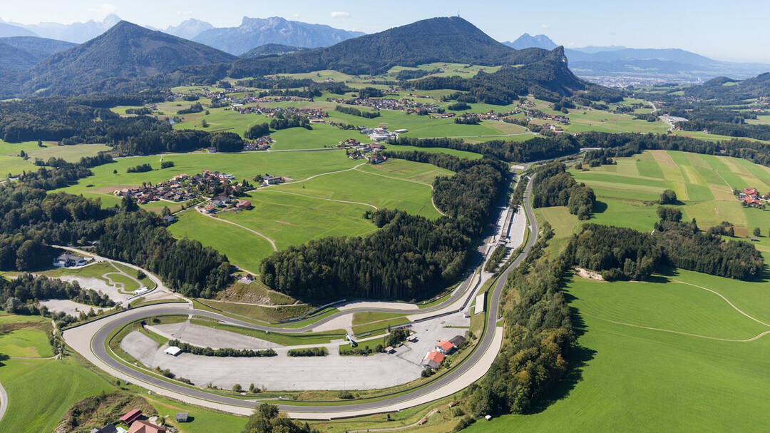 Eine kurvenreiche Rennstrecke, umgeben von grünen Feldern, Bäumen, Bergen und verstreuten Häusern auf dem Lande.