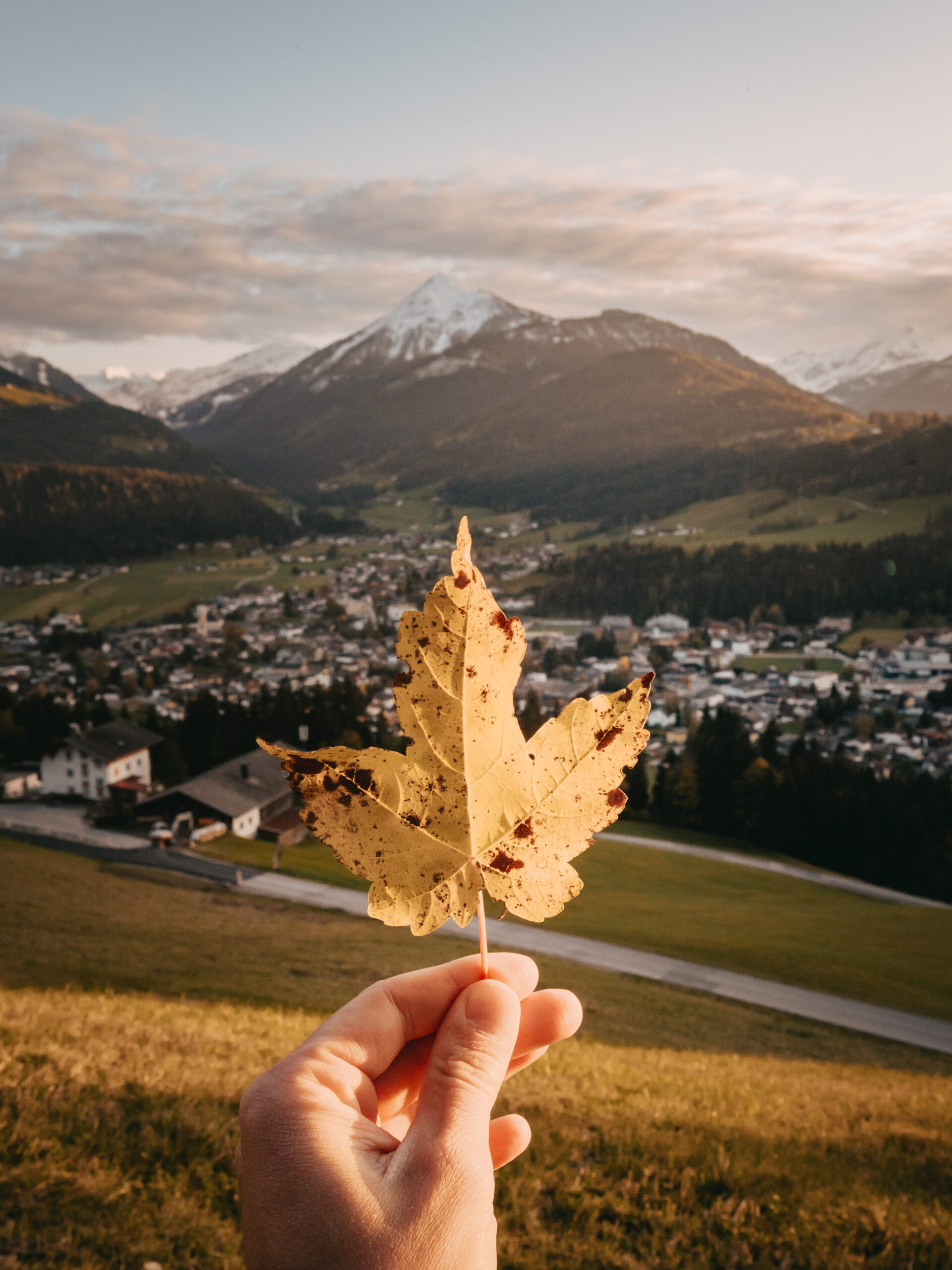 Eine Hand hält ein Blatt mit einem Bergdorf und schneebedeckten Gipfeln im Hintergrund bei Sonnenuntergang.