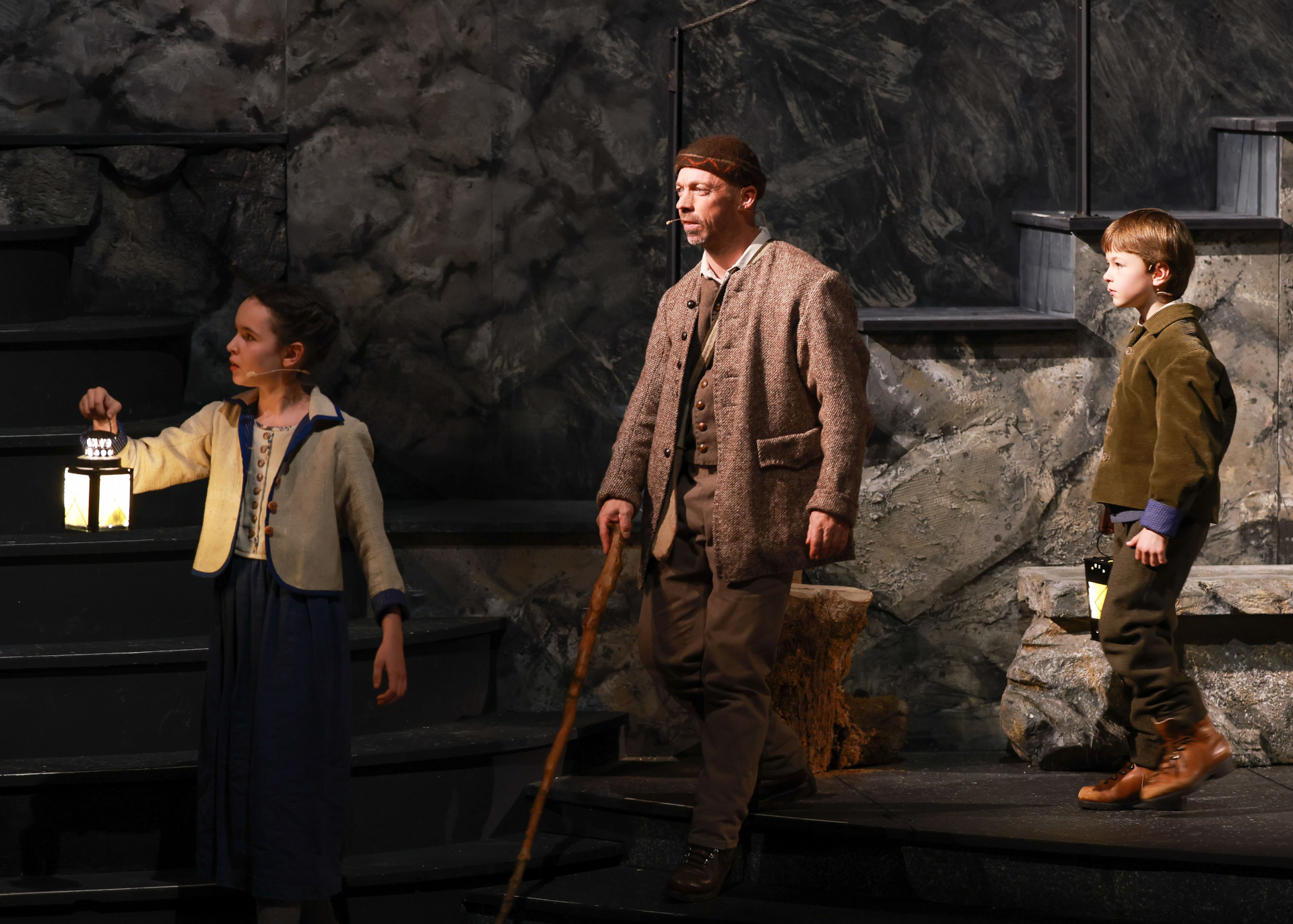 Drei Personen in historischen Kost&uuml;men halten Laternen und stehen auf einem steinernen B&uuml;hnenbild, das an eine Felsenlandschaft erinnert.