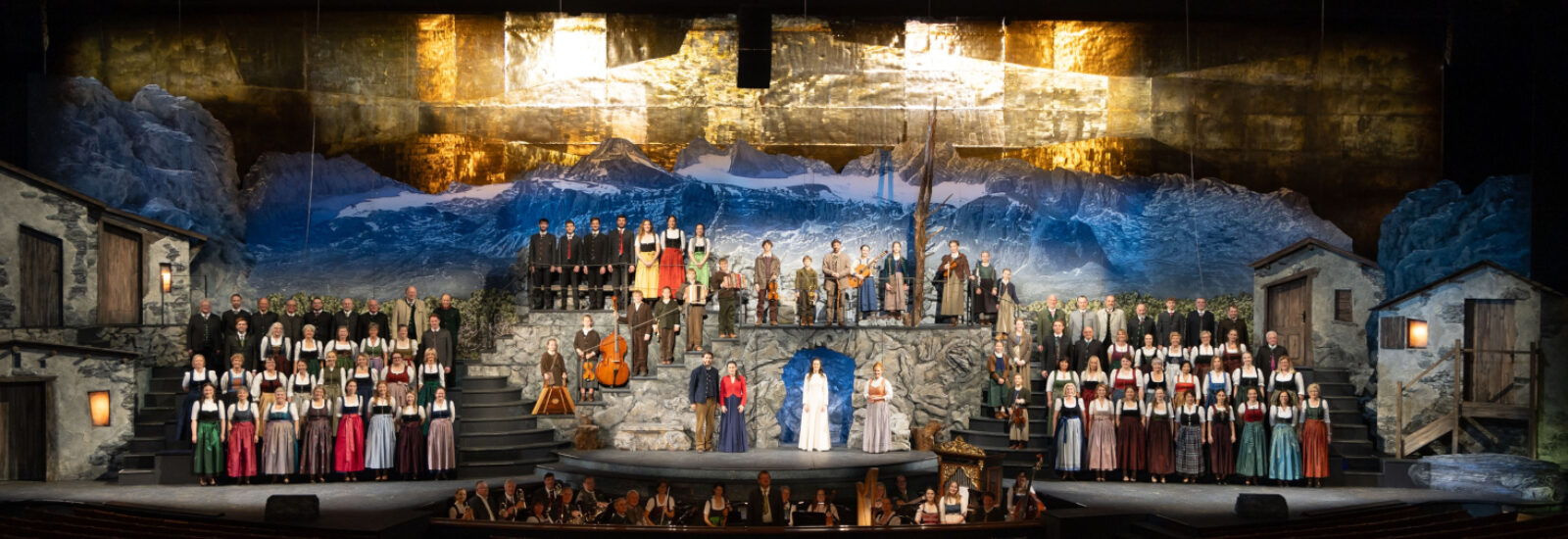 Ein großer Chor in österreichischer Tracht tritt auf einer dramatischen Alpendorfbühne mit Bergkulisse auf.