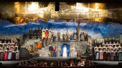 Ein großer Chor in österreichischer Tracht tritt auf einer dramatischen Alpendorfbühne mit Bergkulisse auf.